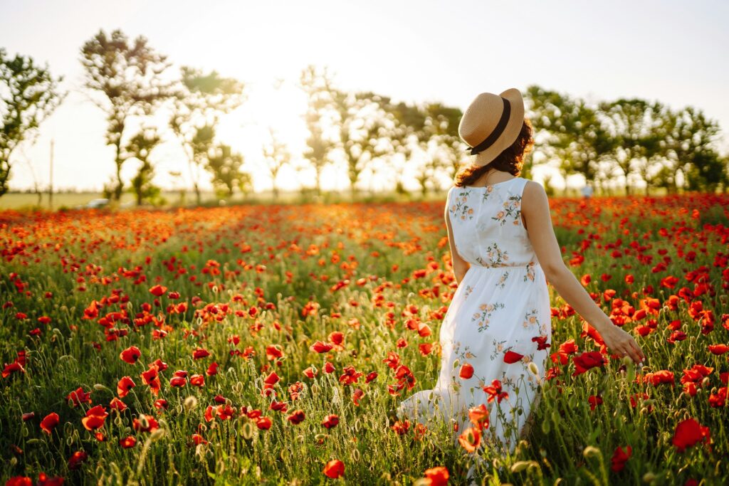 Das Bild zeigt eine Frau, die durch ein weites Mohnblumenfeld spaziert. Sie trägt ein weißes Sommerkleid mit Blumenmuster und einen Strohhut. Die leuchtend roten Mohnblumen erstrecken sich über das ganze Feld, während die untergehende Sonne die Szene in ein warmes, goldenes Licht taucht. Im Hintergrund sind unscharf stehende Bäume zu sehen, die eine natürliche Grenze des Feldes bilden. Die Frau ist von hinten zu sehen und vermittelt ein Gefühl von Freiheit und Gelassenheit in dieser idyllischen Naturkulisse.